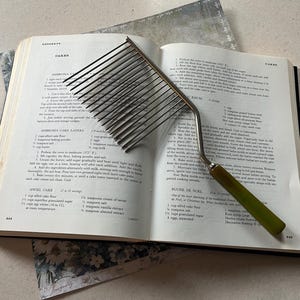 May include: A cake leveler with a green handle rests on an open cookbook. The leveler has a series of thin, parallel blades for slicing cakes evenly. The cookbook is open to a page with cake recipes.
