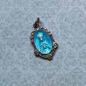May include: A blue enamel pendant with a gold-toned frame and a depiction of a crowned figure. The pendant features the text "YOUR DAUGHTER" in a circular frame.