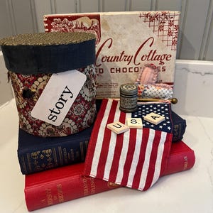May include: A collection of items including a patterned cylindrical box with a tag reading story, a vintage Country Cottage chocolates box, a small American flag, three Scrabble tiles spelling USA, a small metal tin, and two hardcover books in red and blue.