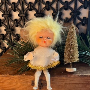 May include: A vintage Christmas angel doll with a yellow fluffy hair, wearing a white dress with gold trim. A small gold Christmas tree is next to the doll. The doll is on a wooden surface.