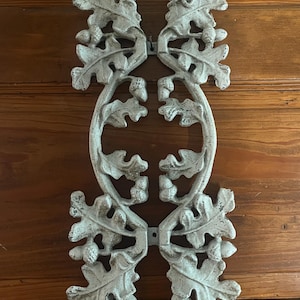 May include: A white, distressed-finish metal wall plaque featuring a symmetrical design of oak leaves and acorns. The elongated, vertical piece has an open, oval-shaped center and two small mounting holes at the top and bottom for attachment.