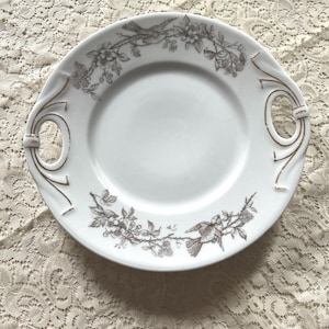 May include: White porcelain serving plate with gold trim and a black and white floral design featuring birds. The plate has two handles.