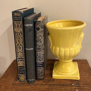 May include: A yellow ceramic urn with a square base and two handles stands next to three antique books with dark covers and gold lettering. The books are arranged vertically, with the urn on the right.