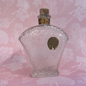 May include: Clear glass perfume bottle with a decorative design. The bottle has a cork stopper and a gold-colored band around the neck. A gold-colored emblem is on the front of the bottle. The bottle is on a pink background.