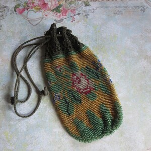 May include: A drawstring pouch crafted from small glass beads in shades of gold, green, and pink, featuring a floral pattern. The top of the pouch is finished with a dark green crocheted edge and includes a matching dark green cord drawstring.