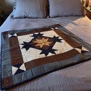 May include: A square quilt featuring a central tan star surrounded by dark blue star shapes on a cream background with small dark dots. The quilt has a wide brown border and an outer edge of blue and black plaid fabric with corner accents.