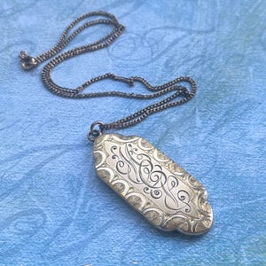 May include: Silver-toned rectangular pendant necklace with an ornate, engraved design. The pendant features a decorative border and a central monogram. The chain is a matching silver color. The necklace is displayed on a blue surface.