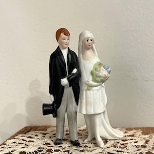 May include: A vintage porcelain wedding cake topper featuring a bride and groom. The groom wears a black tuxedo with a top hat, and the bride is in a white dress and veil, holding a bouquet of flowers. The figures are painted with delicate details.