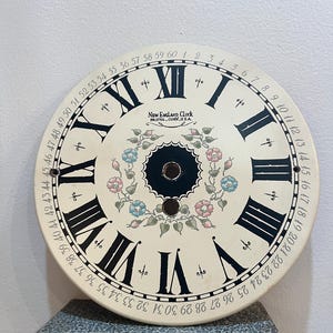 May include: Cream-colored clock face with black Roman numerals and minute markers. The center features a floral design with pink and blue flowers. The text "NEW ENGLAND CLOCK BRISTOL, CONN. U.S.A." is visible. The clock face is approximately 12 inches in diameter.