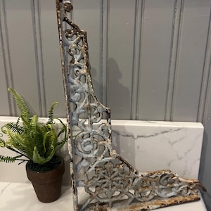 May include: An L-shaped metal shelf bracket featuring an intricate openwork scroll design. The bracket has a distressed finish with visible patches of light blue paint over a dark, rusted brown metal surface, showing signs of age and wear.
