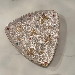 May include: A white triangular ceramic dish with a textured surface and gold leaf accents. The dish is decorated with a floral pattern and small colorful dots.
