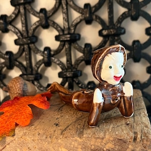 May include: A vintage ceramic figurine of a child in a brown outfit, with a white face, green eyes, and red lips. The figurine is lying down on a wooden surface, with an orange leaf and an acorn nearby. The background features a decorative metalwork.