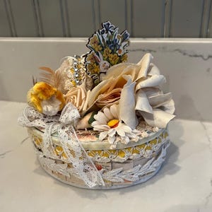 May include: A round, decorative box with a floral arrangement on top. The box is adorned with lace, ribbons, and paper flowers in cream, yellow, and white. A small sticker of a bunny and flowers is attached to the top.