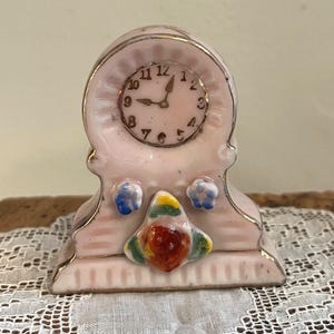 May include: A small, pale pink ceramic mantel clock featuring gold-tone trim, a round clock face with black numerals and hands, and a decorative molded floral accent in red, yellow, and green at the base, flanked by two small blue flowers.