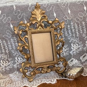 May include: A gold-toned ornate picture frame with a decorative design. The frame has a beaded border and a rectangular opening for a photo.