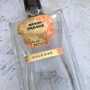 May include: An empty, clear glass cologne bottle with a black cap. The label reads "Dress Parade" in gold lettering with red and gold decorative accents. The word "Cologne" is also visible on a gold label.