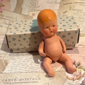 May include: A vintage doll with a peach-colored body and orange hair sits in front of a box with blue heart patterns. The doll has a painted face with rosy cheeks and a small mouth. The box is light blue and white.