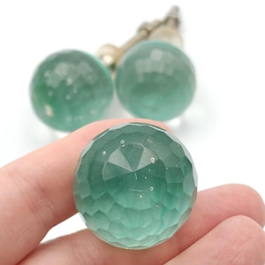 May include: A faceted, teal-colored crystal sphere is held in a hand, with two similar spheres and a silver-toned metal chain in the background. The sphere has a geometric pattern of many small, flat surfaces.