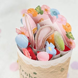 May include: A decorative Easter-themed paper basket filled with colorful paper eggs, flowers, and bunny ears. The basket is made of woven paper and has a lace trim. The arrangement includes pink, blue, orange, and green elements.
