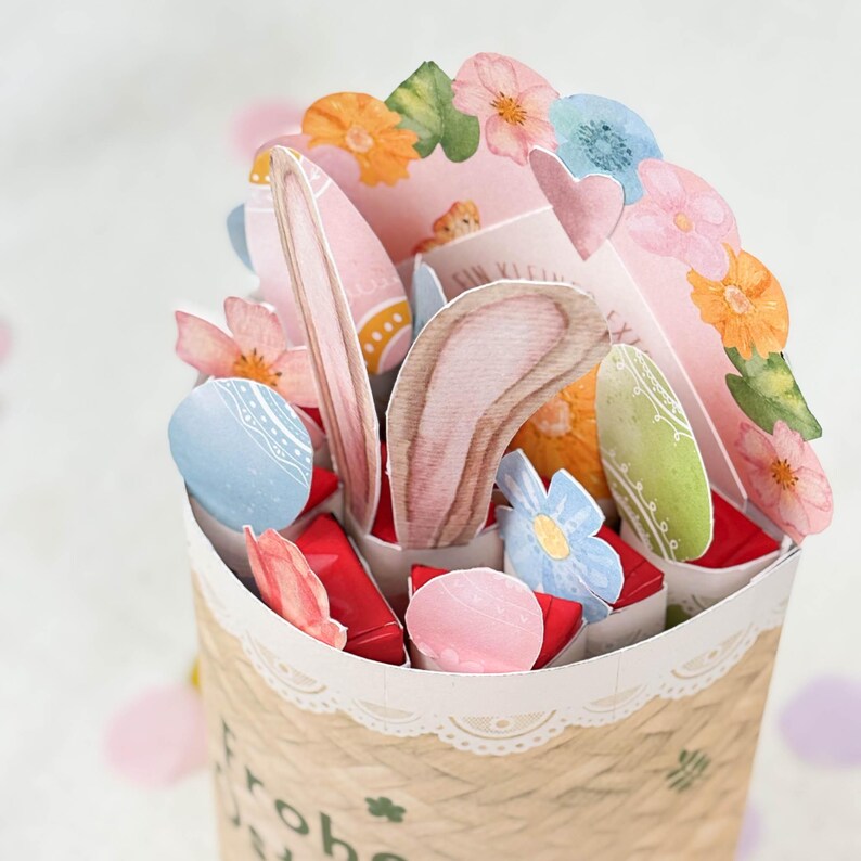 May include: A decorative Easter-themed paper basket filled with colorful paper eggs, flowers, and bunny ears. The basket is made of woven paper and has a lace trim. The arrangement includes pink, blue, orange, and green elements.