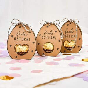 Puede incluir: Tres cajas de regalo marrones en forma de huevo con el texto alem&aacute;n "Frohe Ostern!" y un recorte en forma de coraz&oacute;n que revela un chocolate dorado. Cada caja tiene una asa de cuerda y est&aacute; decorada con estrellas. Confeti esparcido.