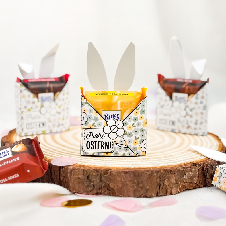 Puede incluir: Barras de chocolate de Pascua en cajas decorativas con orejas de conejo. Las cajas est&aacute;n decoradas con motivos florales y el texto "Frohe OSTERN!". Se exhiben sobre una rodaja de madera con confeti.