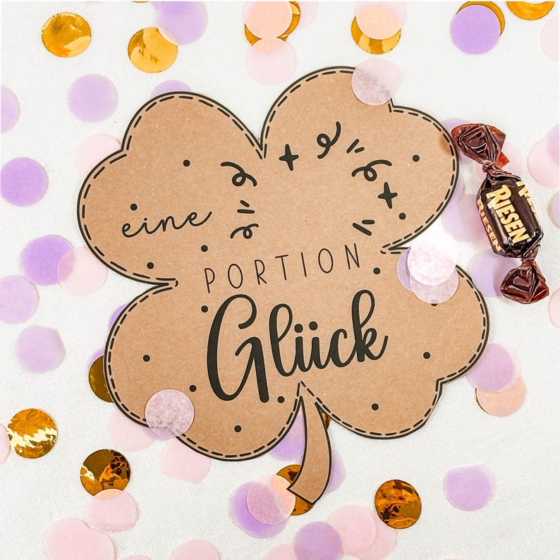 May include: A brown, four-leaf clover-shaped card with the German words "eine Portion Gl&uuml;ck" (a portion of luck) in black script. The card is surrounded by confetti in gold, pink, and purple. A wrapped candy is on the right.