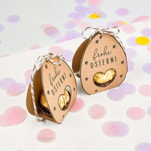 Puede incluir: Dos cajas de regalo marrones con forma de huevo de Pascua con una ventana en forma de coraz&oacute;n que revela un chocolate dorado. Cada caja tiene el texto "Frohe OSTERN!" y detalles de estrellas. Las cajas est&aacute;n atadas con hilo y se encuentran entre confeti pastel.