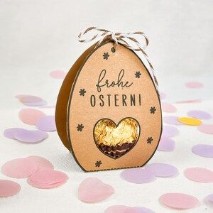 Puede incluir: Una caja de regalo marr&oacute;n en forma de huevo de Pascua con las palabras "Frohe OSTERN!" y una ventana en forma de coraz&oacute;n que revela un chocolate envuelto en oro. La caja est&aacute; decorada con peque&ntilde;as formas de estrella y atada con un lazo de cuerda. Confeti rosa, morado y amarillo rodea la caja.