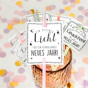 May include: A clear glass with a textured surface holds several paper signs with text and star designs. The signs are attached to straws of various colors. The main sign reads "EIN KLEINES Licht FÜR EIN STRAHLENDES NEUES JAHR!"
