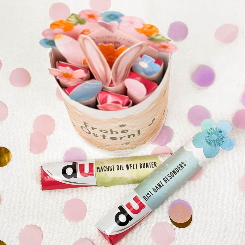 May include: An Easter-themed gift basket with a bunny ear design, filled with colorful candies and treats. Two wrapped candies with the word "du" are visible, along with confetti.
