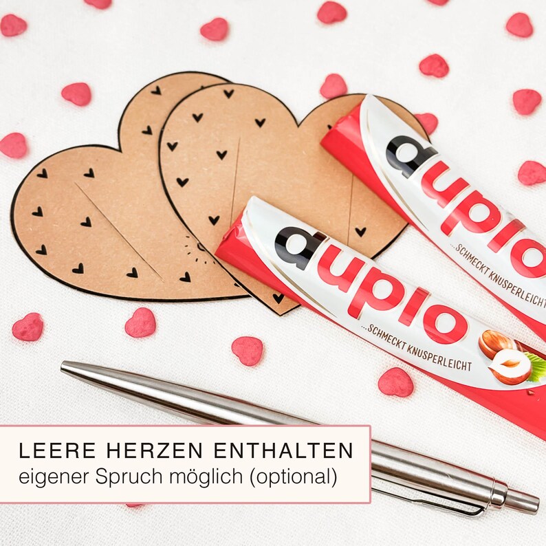 May include: Two brown heart-shaped cutouts with black heart details, alongside two red and white Duplo candy bars. A silver pen and pink heart-shaped candies are scattered around the items. Text on the image reads: "LEERE HERZEN ENTHALTEN eigener Spruch m&ouml;glich (optional)".