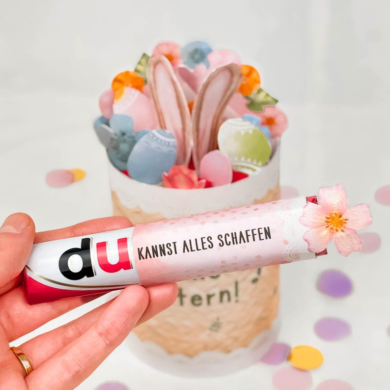 May include: A decorative Easter-themed gift box with a paper-cut design, filled with colorful eggs, flowers, and bunny ears. A tube with the text "KANNST ALLES SCHAFFEN" is held in front of the box. The tube is decorated with a pink flower.