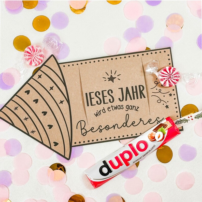 Puede incluir: Una escena festiva con un gorro de fiesta de papel marr&oacute;n y una tarjeta que dice "IESES JAHR wird etwas ganz Besonderes". Una barra de chocolate Duplo y caramelos envueltos est&aacute;n esparcidos entre confeti dorado, rosa y morado.