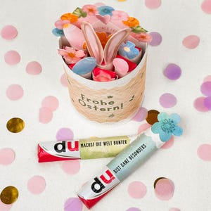 May include: An Easter-themed gift basket with a floral arrangement, bunny ears, and the words "Frohe Ostern!" A red and white candy bar with the word "du" and the text "MACHST DIE WELT BUNTER" is next to a blue and white candy bar with the word "du" and the text "BIST GANZ BESONDERS".
