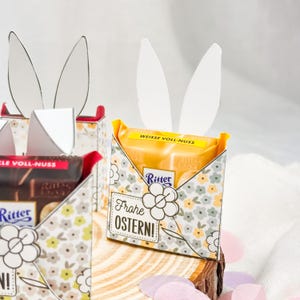 Puede incluir: Cajas de regalo con tem&aacute;tica de Pascua con orejas de conejo blancas. Las cajas est&aacute;n decoradas con un estampado floral y contienen barras de chocolate Ritter. Una caja tiene el texto "Frohe OSTERN!" y "WEISSE VOLL-NUSS".