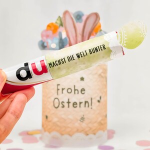 May include: A decorative Easter-themed gift. The image shows a tube with the word "du" and the text "MACHST DIE WELT BUNTER" and "Frohe Ostern!" on it. The tube is held in front of a decorative basket with bunny ears and colorful decorations.