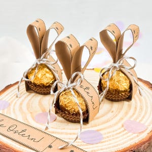 May include: Three Easter-themed chocolate candies wrapped in gold foil and brown paper cups. Each candy is adorned with brown paper bunny ears and tied with twine. The candies are on a wooden slice with decorative confetti.