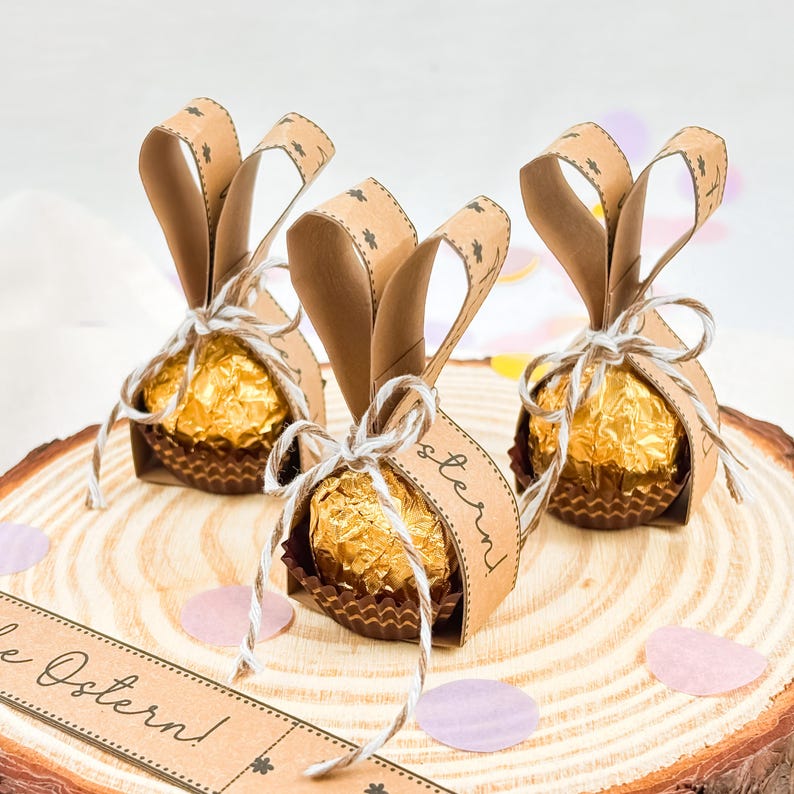 May include: Three Easter-themed chocolate candies wrapped in gold foil and brown paper cups. Each candy is adorned with brown paper bunny ears and tied with twine. The candies are on a wooden slice with decorative confetti.