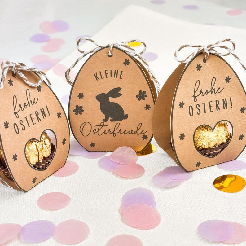 Puede incluir: Tres cajas de regalo marrones con forma de huevo de Pascua con una ventana en forma de coraz&oacute;n. Las cajas tienen las palabras "frohe OSTERN!" y "KLEINE Osterfreude" impresas, junto con una silueta de conejo. Las cajas est&aacute;n atadas con hilo.