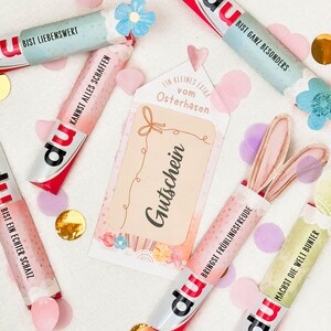 May include: An assortment of colorful candy bars with German text, scattered with confetti and a gift tag that reads "Gutschein". Some candy bars have bunny ears attached. The candy bars have the brand name "du".