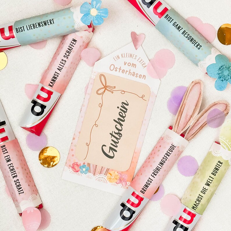 May include: An assortment of colorful candy bars with German text, scattered with confetti and a gift tag that reads "Gutschein". Some candy bars have bunny ears attached. The candy bars have the brand name "du".