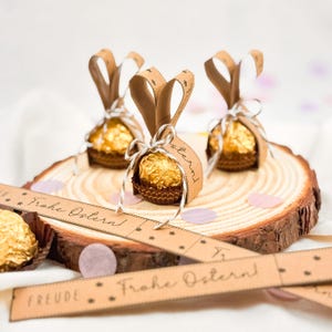 May include: Three Easter-themed chocolate candies wrapped in gold foil and brown paper cups, decorated with brown paper bunny ears and tied with white string. The candies sit on a wooden slice with the text "Frohe Ostern!" on a brown paper ribbon.