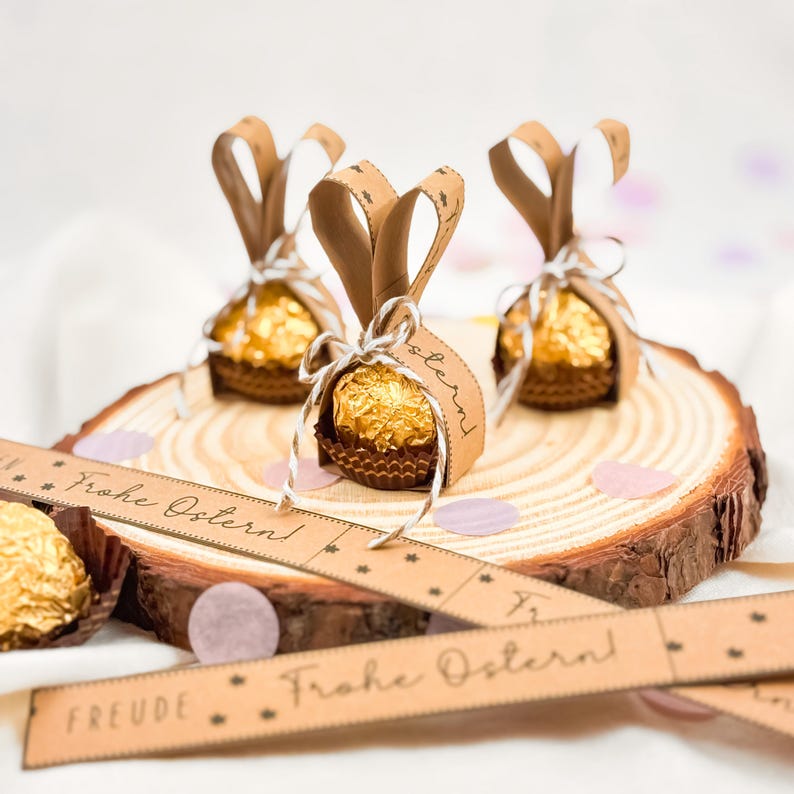 May include: Three Easter-themed chocolate candies wrapped in gold foil and brown paper cups, decorated with brown paper bunny ears and tied with white string. The candies sit on a wooden slice with the text "Frohe Ostern!" on a brown paper ribbon.