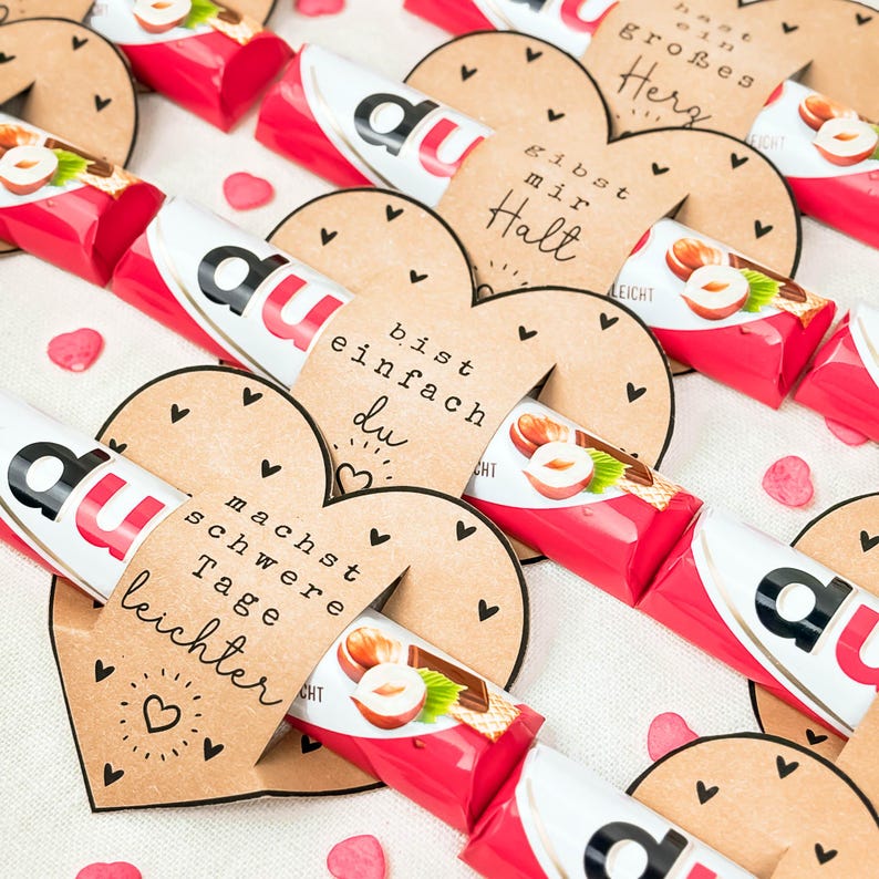 May include: Arrangement of red and white candy bars with hazelnut illustrations, paired with heart-shaped tags. The tags feature German text and small black heart designs, creating a festive and personalized gift.