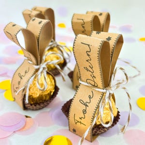 May include: Several Easter-themed gift packages. Each package features a gold-wrapped chocolate candy in a brown paper cup, tied with twine, and adorned with a brown paper ribbon that reads "Frohe Ostern!" in a script font. Confetti in various colors is scattered around.