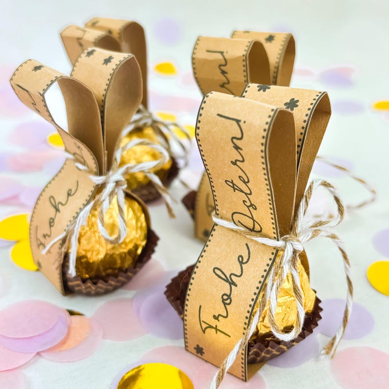 May include: Several Easter-themed gift packages. Each package features a gold-wrapped chocolate candy in a brown paper cup, tied with twine, and adorned with a brown paper ribbon that reads "Frohe Ostern!" in a script font. Confetti in various colors is scattered around.