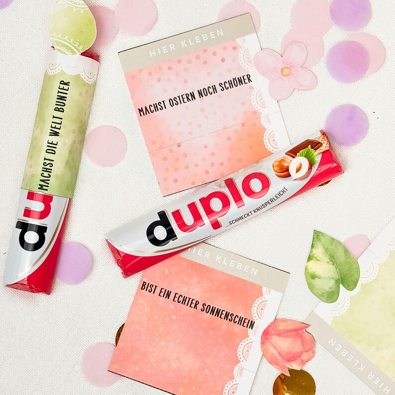 May include: An overhead shot features a Duplo candy bar with hazelnut and wafer filling, and two other candy bars with text. The image includes decorative cards with text, paper flowers, and confetti in pink, purple, and green.