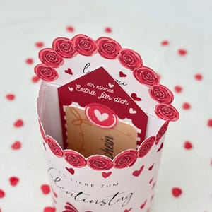 Pu&ograve; includere: Un contenitore regalo di San Valentino con un design floreale rosso e bianco. Il contenitore contiene una carta rossa con il testo "ein kleines Extra f&uuml;r dich" e una carta regalo. Coriandoli a forma di cuore rosso circondano il contenitore.
