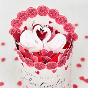 Pu&ograve; includere: Un'opera di carta a tema San Valentino con due cigni bianchi con colli intrecciati, circondati da cuori e rose rosse. Il design include un bordo decorativo di rose rosse e le parole "Alles Liebe zum Valentinstag."